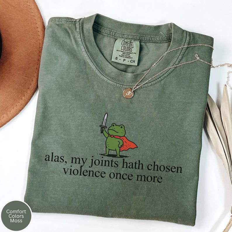 May include: A moss green Comfort Colors t-shirt with a cartoon frog holding a sword and wearing a cape. The text "alas, my joints hath chosen violence once more" is printed below the frog. A gold necklace is draped across the shirt.