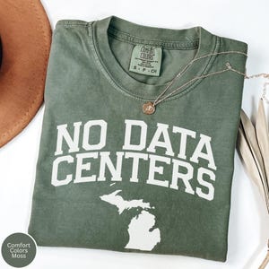 May include: An olive green t-shirt with the text "NO DATA CENTERS" in white block letters and a white outline of the state of Michigan. A gold necklace with a pendant rests on the shirt. The shirt is labelled "Comfort Colors Moss".