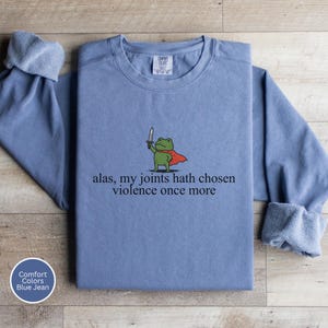 May include: A blue jean-coloured sweatshirt featuring the phrase "alas, my joints hath chosen violence once more." The design includes a cartoon frog in a red cape, holding a sword. The sweatshirt has a crew neck and long sleeves.
