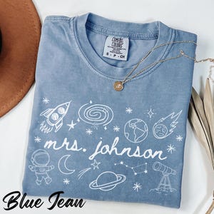 Puede incluir: Camiseta azul vaquero con un gráfico blanco con temática espacial y el texto "mrs. johnson". El diseño incluye un cohete, un astronauta, planetas y estrellas. Un collar dorado con un colgante está sobre la camiseta.