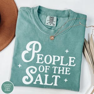 May include: A light green Comfort Colors t-shirt with the text "People of the Salt" in white script. The shirt is folded and has a small tag at the neckline. A gold necklace with a pendant is draped across the shirt.