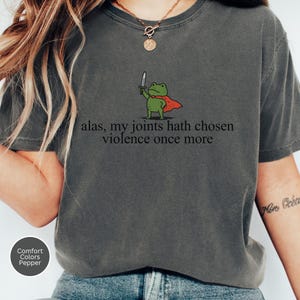 May include: Dark grey t-shirt with a cartoon frog in a red cape and sword. The text "alas, my joints hath chosen violence once more" is printed on the shirt. The shirt is a Comfort Colors Pepper tee.