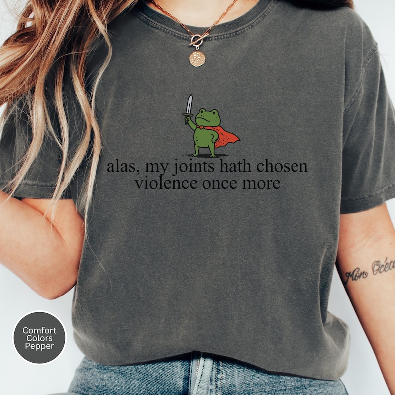 May include: Dark grey t-shirt with a cartoon frog in a red cape and sword. The text "alas, my joints hath chosen violence once more" is printed on the shirt. The shirt is a Comfort Colors Pepper tee.