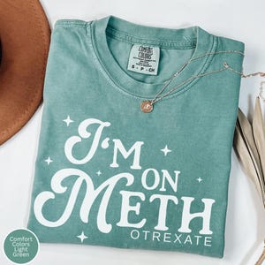 May include: Light green Comfort Colors t-shirt with the text "I'm on Meth Otrexate" in white script. The shirt has a crew neck and short sleeves. A gold necklace with a pendant is draped across the shirt.