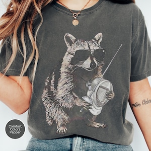 May include: A charcoal grey short-sleeve t-shirt featuring a graphic print of a raccoon wearing black sunglasses and holding a fencing mask and foil. The shirt has a round neckline and a small circular label in the corner reading Comfort Colors Pepper.