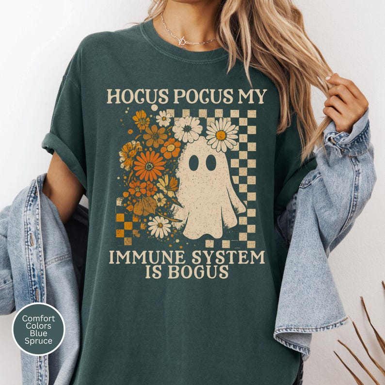 May include: Dark teal t-shirt with the text "Hocus Pocus My Immune System Is Bogus." The shirt features a ghost illustration with flowers and a checkered pattern. The shirt is a comfort colors brand in blue spruce.