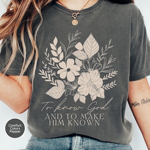 Può includere: T-shirt grigio scuro con un design floreale beige e la scritta "To know God AND TO MAKE HIM KNOWN". La maglietta è una Comfort Colors Pepper.