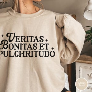 May include: A sand-colored sweatshirt with the words "Veritas Bonitas Et Pulchritudo" in black script. The sweatshirt has a crew neck and long sleeves. The Gildan Sand color is displayed in the bottom right corner.