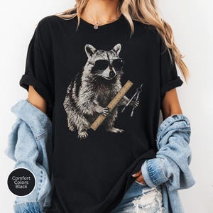 May include: A black t-shirt featuring a graphic print of a raccoon wearing sunglasses. The raccoon is holding a wooden ruler and a metal drafting compass. The shirt has a crew neckline and short sleeves, with a small circular label reading Comfort Colors Black.