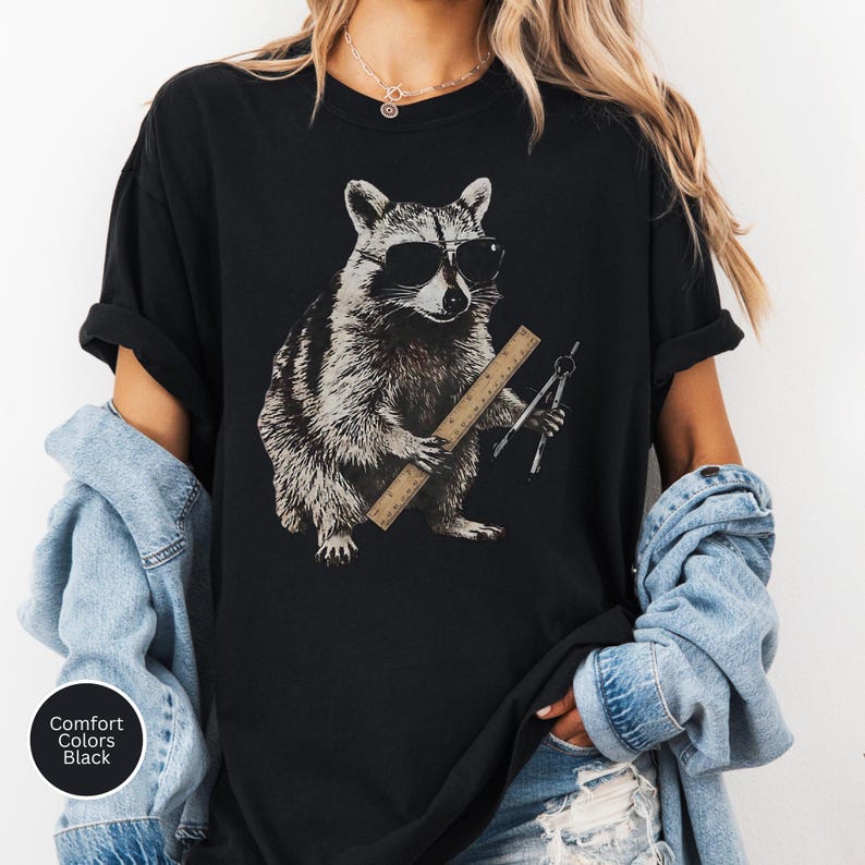 May include: A black t-shirt featuring a graphic print of a raccoon wearing sunglasses. The raccoon is holding a wooden ruler and a metal drafting compass. The shirt has a crew neckline and short sleeves, with a small circular label reading Comfort Colors Black.