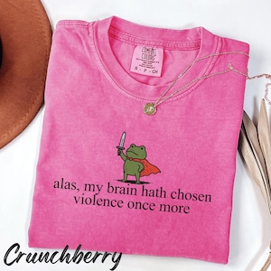 Op de afbeelding: Een roze Comfort Colors t-shirt met de tekst "alas, my brain hath chosen violence once more" en een cartoon kikker met een zwaard. Het shirt heeft ook het woord "Crunchberry" erop gedrukt.