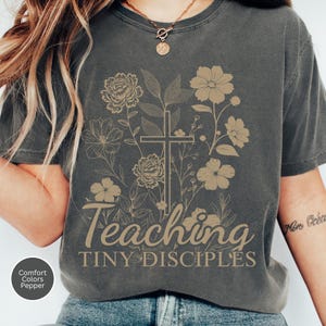 May include: Dark gray t-shirt with a floral design and a cross. The words "Teaching Tiny Disciples" are printed in a light brown color. The shirt is made of a soft, comfortable material.