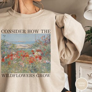 Puede incluir: Sudadera color arena con el texto "CONSIDER HOW THE WILDFLOWERS GROW" encima y debajo de una pintura de paisaje floral. La pintura presenta flores silvestres rojas, blancas y moradas cerca de una masa de agua.
