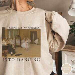 Puede incluir: Sudadera color arena con una imagen impresa de una escena de ballet y el texto "HE TURNS MY MOURNING INTO DANCING". La imagen presenta bailarinas con tutús blancos y un hombre tocando el violín. La sudadera es color arena Gildan.