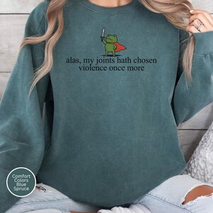 May include: Blue spruce sweatshirt featuring the phrase "alas, my joints hath chosen violence once more." A cartoon frog in a red cape and holding a sword is printed above the text. The sweatshirt has a crew neck and long sleeves.