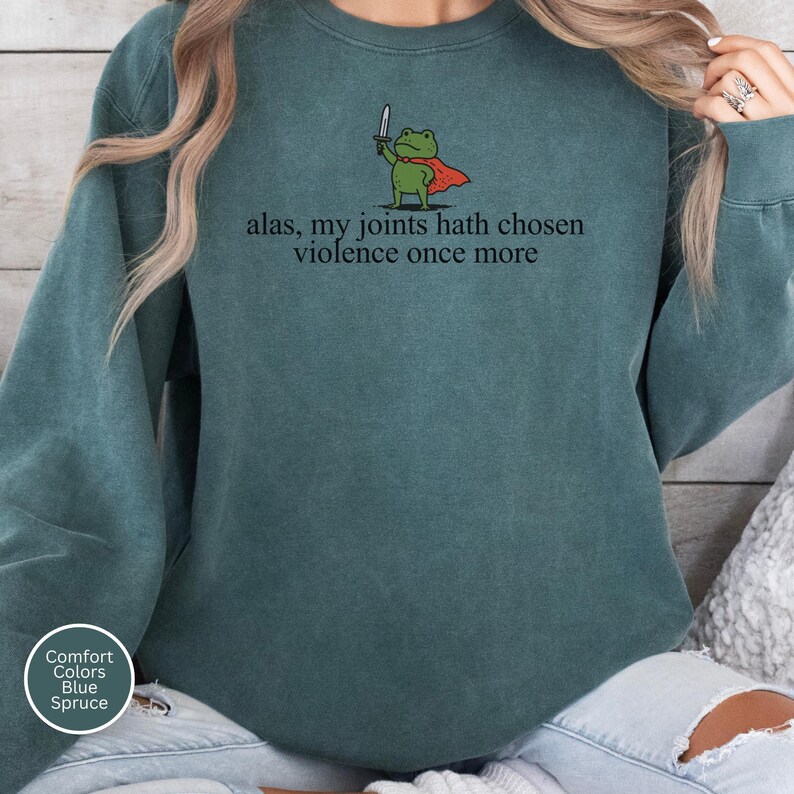 May include: Blue spruce sweatshirt featuring the phrase "alas, my joints hath chosen violence once more." A cartoon frog in a red cape and holding a sword is printed above the text. The sweatshirt has a crew neck and long sleeves.