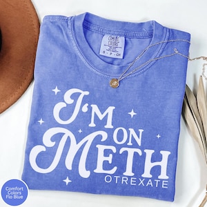 May include: A blue Comfort Colors t-shirt with the text "I'm on Meth Otrexate" in white. The shirt has a crew neck and short sleeves. A gold necklace with a pendant is draped across the shirt.