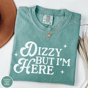 Puede incluir: Una camiseta verde claro Comfort Colors con el texto "Dizzy But I'm Here" en escritura blanca. Un collar dorado con un colgante descansa sobre la camiseta. La camiseta está doblada y colocada sobre una superficie neutra.