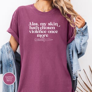 May include: Berry-colored t-shirt with rolled sleeves. The shirt features the text "Alas, my skin hath chosen violence once more" and a knife graphic in white. The shirt is part of the Comfort Colors collection.