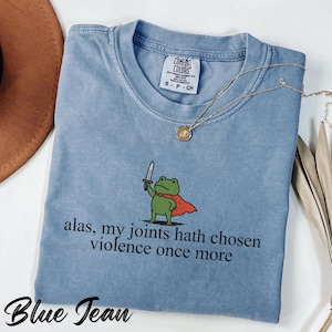 May include: A blue Comfort Colors t-shirt featuring a cartoon frog in a red cape, holding a sword, with the text "alas, my joints hath chosen violence once more". A gold necklace is draped across the shirt. The shirt is labelled "Blue Jean".