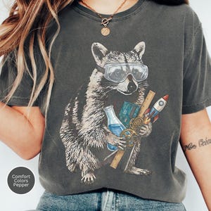 May include: A charcoal grey t-shirt featuring a graphic of a raccoon wearing safety goggles. The raccoon holds a blue liquid-filled beaker, a circuit board, a wooden ruler, and a small rocket ship. The design includes metallic gold gears.