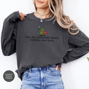 May include: Dark grey sweatshirt featuring the text "alas, my joints hath chosen violence once more." A cartoon frog in a cape with a sword is printed above the text. The sweatshirt is a Comfort Colors brand in the colour Pepper.