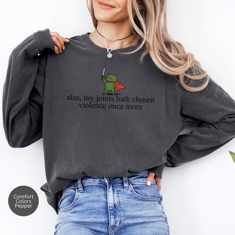 May include: Dark grey sweatshirt featuring the text "alas, my joints hath chosen violence once more." A cartoon frog in a cape with a sword is printed above the text. The sweatshirt is a Comfort Colors brand in the colour Pepper.