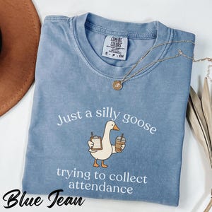 以下が含まれることがあります： 「Just a silly goose trying to collect attendance」の文字が入ったブルーデニムカラーのTシャツ。クリップボードとコーヒーを持った漫画のガチョウが描かれています。金のネックレスがシャツの上にかけられています。