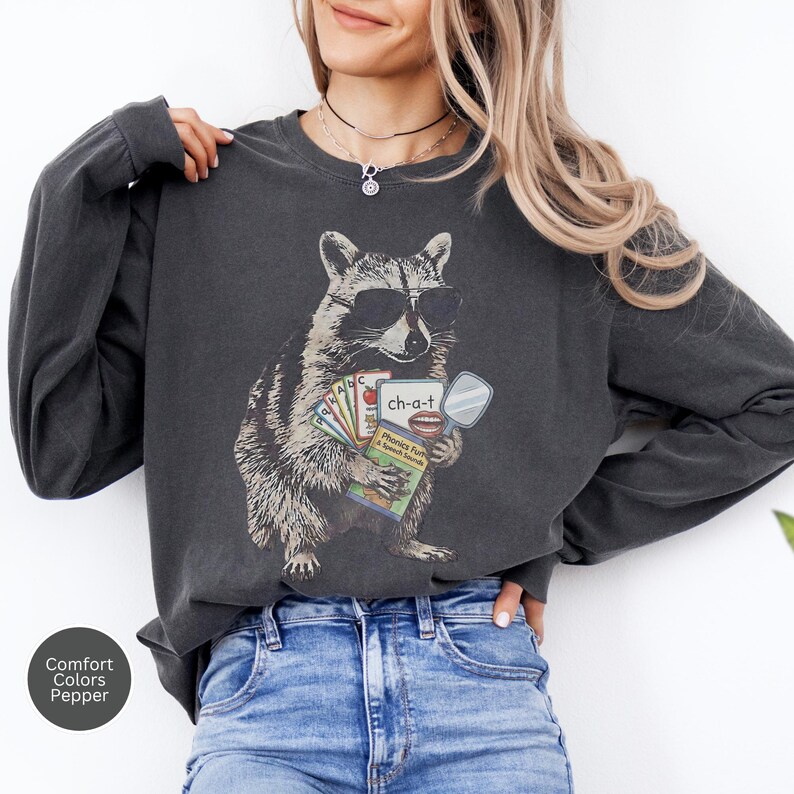 May include: A charcoal gray long-sleeve sweatshirt featuring a graphic of a raccoon wearing sunglasses. The raccoon holds a set of colorful flashcards and a book labeled Phonics Fun & Speech Sounds with the text ch-a-t and an illustration of lips.