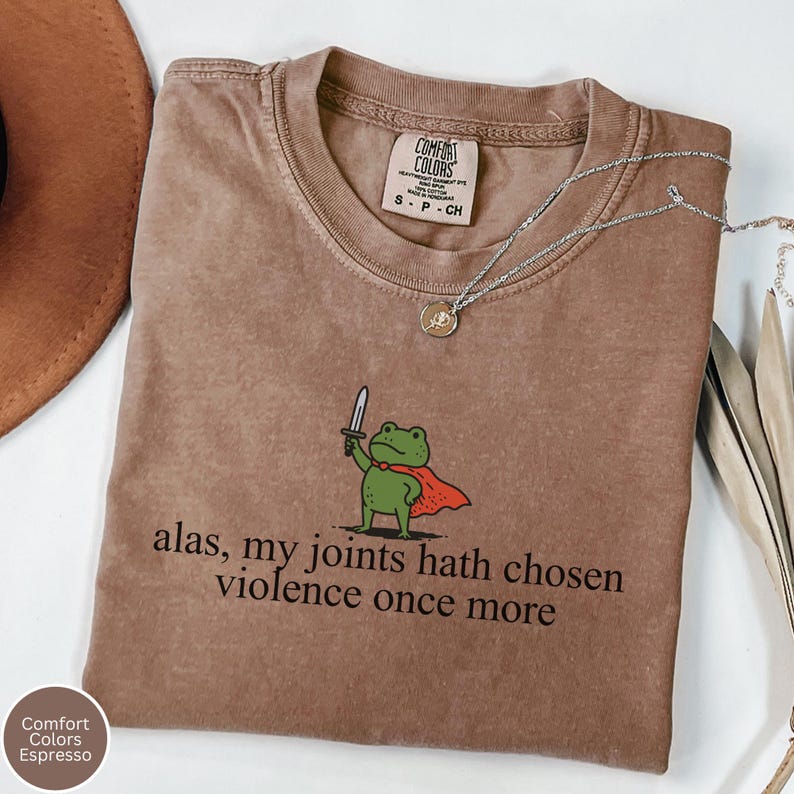 May include: Espresso-coloured Comfort Colors t-shirt featuring a frog graphic with a sword and cape, and the text "alas, my joints hath chosen violence once more." A silver necklace with a pendant is draped on the shirt.