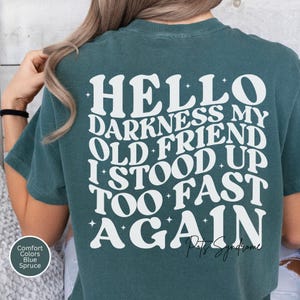 May include: A spruce-colored t-shirt with the text "HELLO DARKNESS MY OLD FRIEND I STOOD UP TOO FAST AGAIN" in white, stylized lettering. The shirt has a vintage, slightly faded look. The text is centered on the back of the shirt.