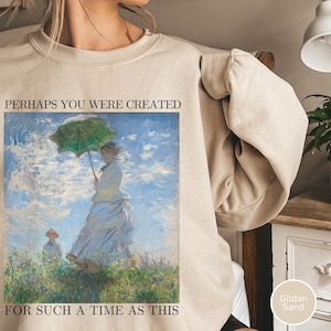Puede incluir: Sudadera color arena con una pintura estilo Monet en la parte delantera. La pintura representa a una mujer con un paraguas verde y un niño en un campo. El texto en la sudadera dice: "PERHAPS YOU WERE CREATED FOR SUCH A TIME AS THIS".