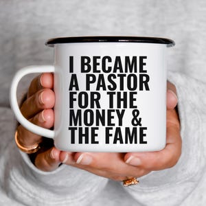 Op de afbeelding: Witte keramische mok met een zwarte rand en oor, met de tekst "I BECAME A PASTOR FOR THE MONEY & THE FAME" in vette zwarte letters. De mok wordt in de handen van een persoon gehouden.