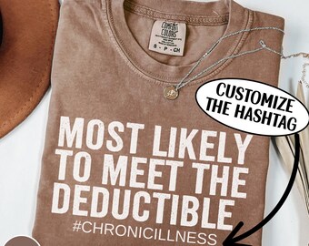 May include: Espresso-colored t-shirt with the text "MOST LIKELY TO MEET THE DEDUCTIBLE" and the hashtag "#CHRONICILLNESS" in white. The shirt has a crew neck and short sleeves. A silver necklace with a pendant is draped across the shirt.