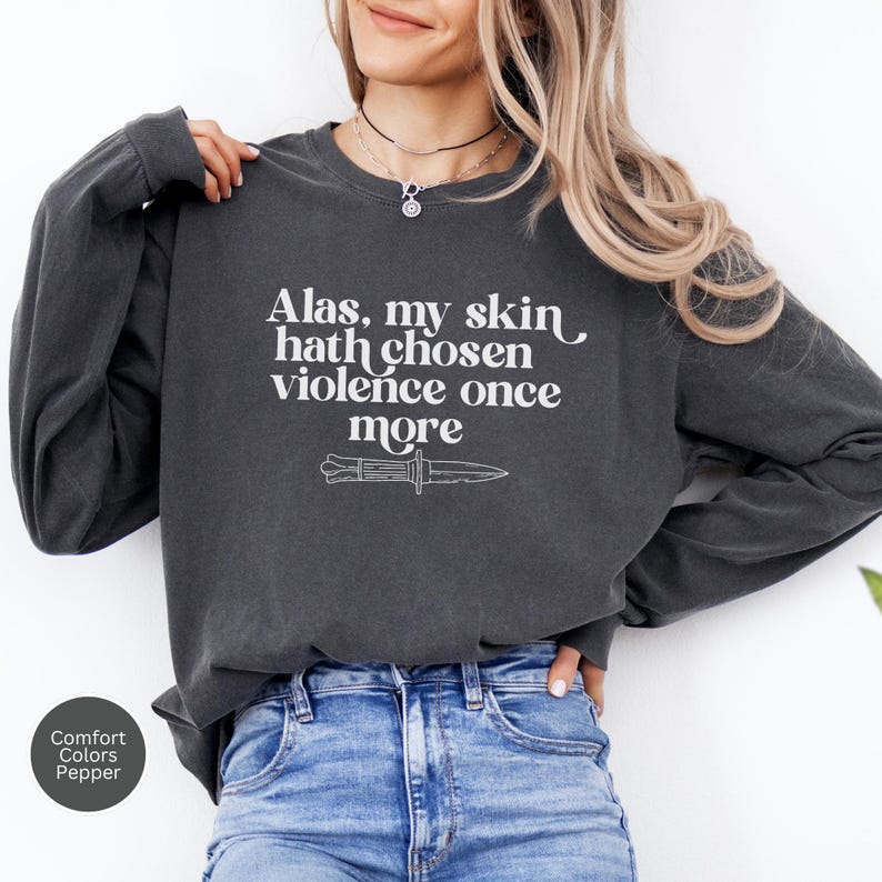 May include: Dark grey long-sleeve sweatshirt with the text "Alas, my skin hath chosen violence once more" and a knife graphic in white. The sweatshirt is a Comfort Colors brand in the color Pepper.