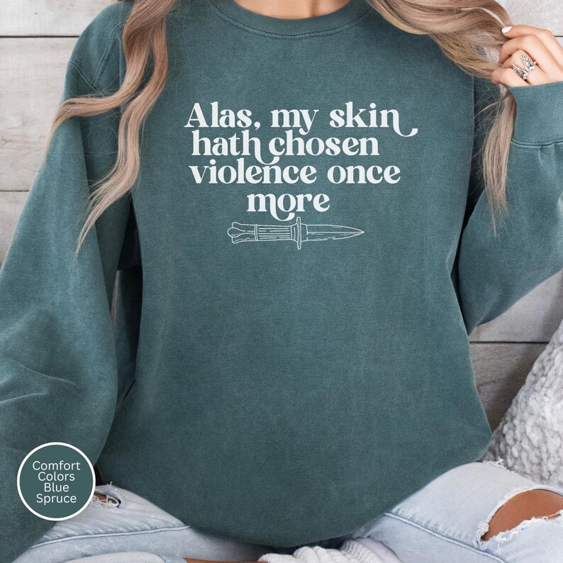 May include: A teal-colored sweatshirt with the text "Alas, my skin hath chosen violence once more" in white, along with a white illustration of a knife. The sweatshirt has a crew neck and long sleeves.