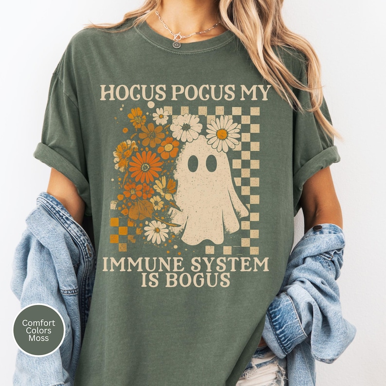 May include: Olive green t-shirt with the text "Hocus Pocus My Immune System Is Bogus." The shirt features a ghost graphic with a floral design and a checkered background. The shirt is made of a soft, comfortable material.
