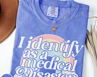 Peut inclure: Un t-shirt bleu Comfort Colors avec le texte "I identify as a medical Disaster" sur un arc-en-ciel. Un collier en or avec un pendentif est drapé sur le t-shirt. Le t-shirt est de taille S.