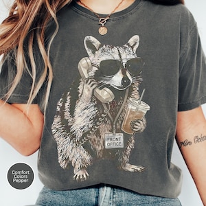Pode incluir: Uma t-shirt cinzento-carvão com um gráfico de um guaxinim a usar óculos de sol pretos, a segurar um auscultador de telefone ao ouvido e um copo de plástico com palhinha. Um crachá com a etiqueta FRONT OFFICE está pendurado ao pescoço.