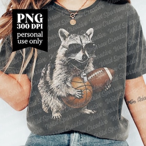 May include: A charcoal gray t-shirt featuring a graphic illustration of a raccoon wearing sunglasses and a whistle around its neck. The raccoon is holding a basketball in its paws, with a brown football positioned next to it on the shirt front.