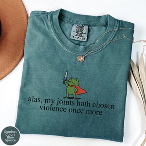 May include: Blue Spruce Comfort Colors t-shirt featuring a cartoon frog with a sword and cape, with the text "alas, my joints hath chosen violence once more". A gold necklace is draped across the shirt.