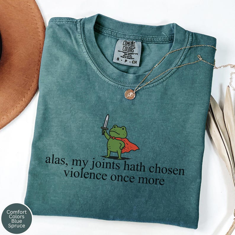 May include: Blue Spruce Comfort Colors t-shirt featuring a cartoon frog with a sword and cape, with the text "alas, my joints hath chosen violence once more". A gold necklace is draped across the shirt.