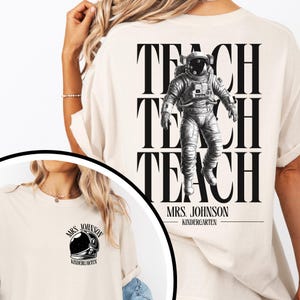 Puede incluir: Una camiseta color crema con un gráfico negro de un astronauta flotando sobre el texto repetido TEACH TEACH TEACH, con MRS. JOHNSON KINDERGARTEN debajo. El área del pecho muestra un gráfico más pequeño de un casco de astronauta negro.