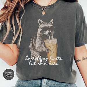 Peut inclure: Un t-shirt gris anthracite avec un motif de raton laveur portant des lunettes de soleil noires et tenant un gobelet en plastique de café glacé. Le texte sous le raton laveur indique, Everything hurts but I'm here. T-shirt à manches courtes.