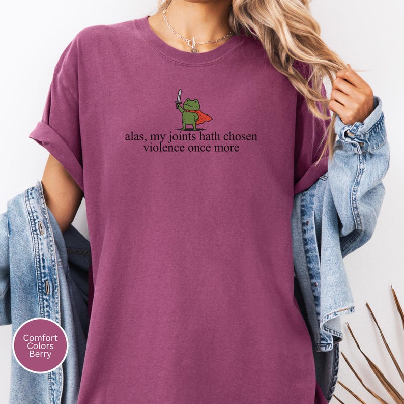 May include: Berry-coloured t-shirt with a cartoon frog holding a knife and wearing a cape. The text "alas, my joints hath chosen violence once more" is printed below the frog. The shirt is a Comfort Colors brand.