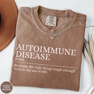 May include: Espresso-colored t-shirt with the text "AUTOIMMUNE DISEASE" in white, along with the phrase "Because the only thing tough enough to kick my ass is me." A silver necklace with a pendant rests on the shirt.