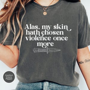 May include: Dark grey t-shirt with the text "Alas, my skin hath chosen violence once more" and a knife illustration. The shirt is a Comfort Colors Pepper tee.