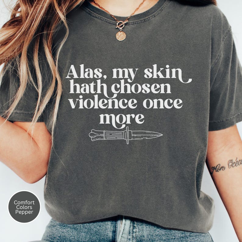 May include: Dark grey t-shirt with the text "Alas, my skin hath chosen violence once more" and a knife illustration. The shirt is a Comfort Colors Pepper tee.