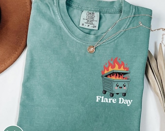 Peut inclure: T-shirt vert clair avec un dessin d'une poubelle souriante en feu et le texte "Flare Day" en dessous. Le t-shirt a un col rond et des manches courtes. Un collier doré est drapé sur le t-shirt.