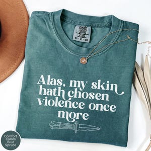 May include: Blue Spruce Comfort Colors t-shirt with the text "Alas, my skin hath chosen violence once more" and a knife graphic. A gold necklace with a pendant is draped on the shirt. The shirt is folded on a white surface.